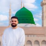 A bearded man smiling in front of the iconic green dome of Al-Masjid an-Nabawi, Medina.